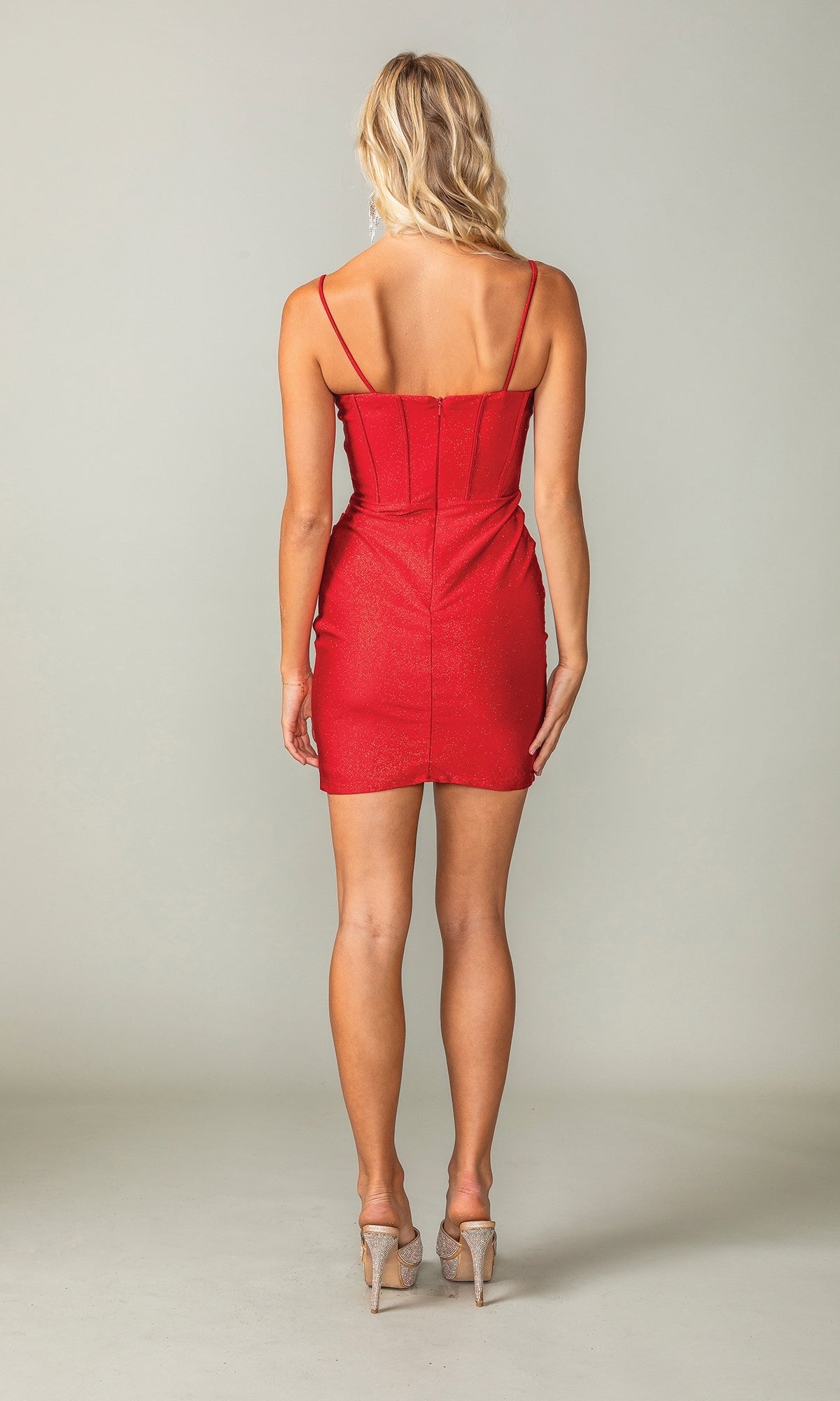 A woman with blonde hair stands facing away against a plain background wearing the Cowl-Neck Short Glitter Homecoming Dress 3396—a fitted, sleeveless red dress—paired with beige high heels.